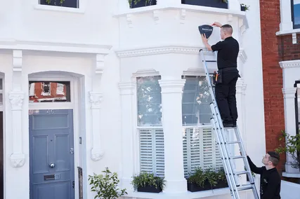 Two workers on a ladder install a Banham security alarm on a white house's exterior, near a bay window with planter boxes, on a sunny day.