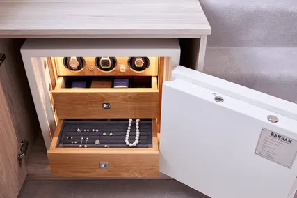 Open safe with wooden drawers containing watches, necklaces, and jewellery, set within a light wooden cabinet.