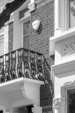 Black and white image of a brick townhouse facade with ornate wrought iron balcony railing and detailed architectural moldings.