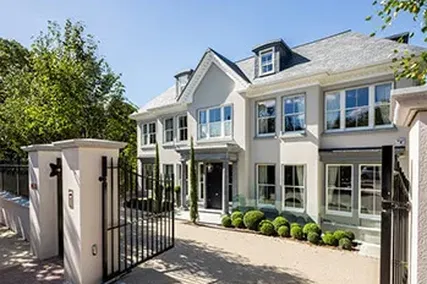 Luxurious two-story house with large windows, a gated entrance, manicured shrubs, and a steep roof under a clear blue sky.