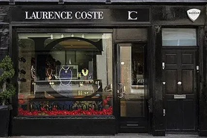Storefront of "Laurence Coste" displaying elegant jewellery in a window, with a sleek black facade and a security badge on the top right.
