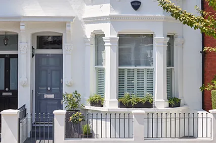 White townhouse exterior with bay window, potted plants, grey bespoke door, and small black fence. Adjacent red-brick building partially visible.