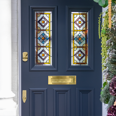 Bespoke Banham Blue door with stained glass panels featuring geometric patterns. Gold hardware and mail slot. Greenery visible on the side.