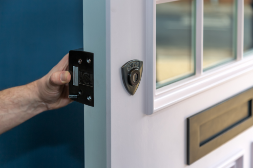 An engineer installing a  lock on a white door with a mail slot and window panels, against a blue wall background.