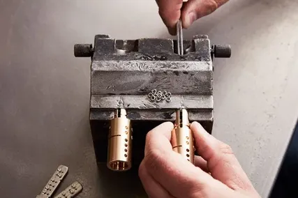Hands working on a metal device, adjusting components with a tool. Two brass cylinders are visible at the front.
