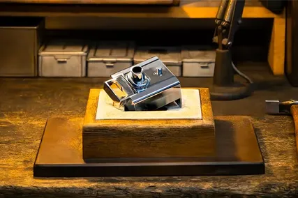 A shiny chrome premium lock mounted on a wooden base, placed on a rustic wooden table with small boxes and tools in the background.