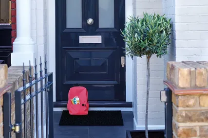 Red backpack on a doormat in front of a luxury black door with a letterbox, beside a small potted tree and brick wall.