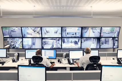 Two security personnel monitoring multiple surveillance screens in a modern control room with desks and computer monitors.