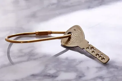 A brass security key with a circular head and several holes along the shaft, attached to a gold keyring, on a marble surface.