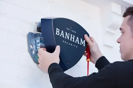 Person installing a Banham security alarm on a white brick wall, holding tools to secure the device.