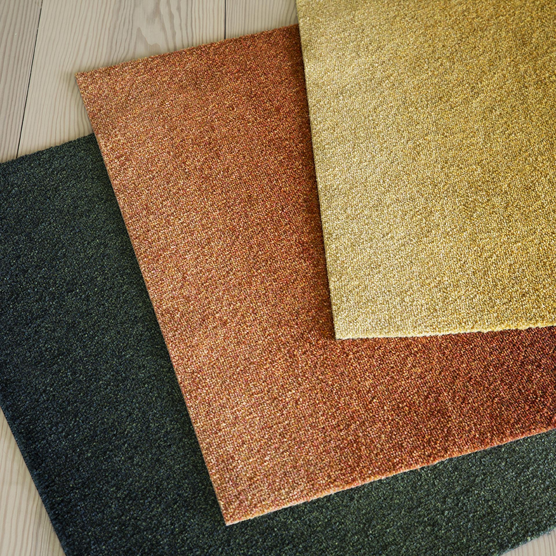 Three doormat samples from Heymats Dis Collection in green, orange, and yellow are layered on a wooden floor, showcasing textured surfaces and warm tones.