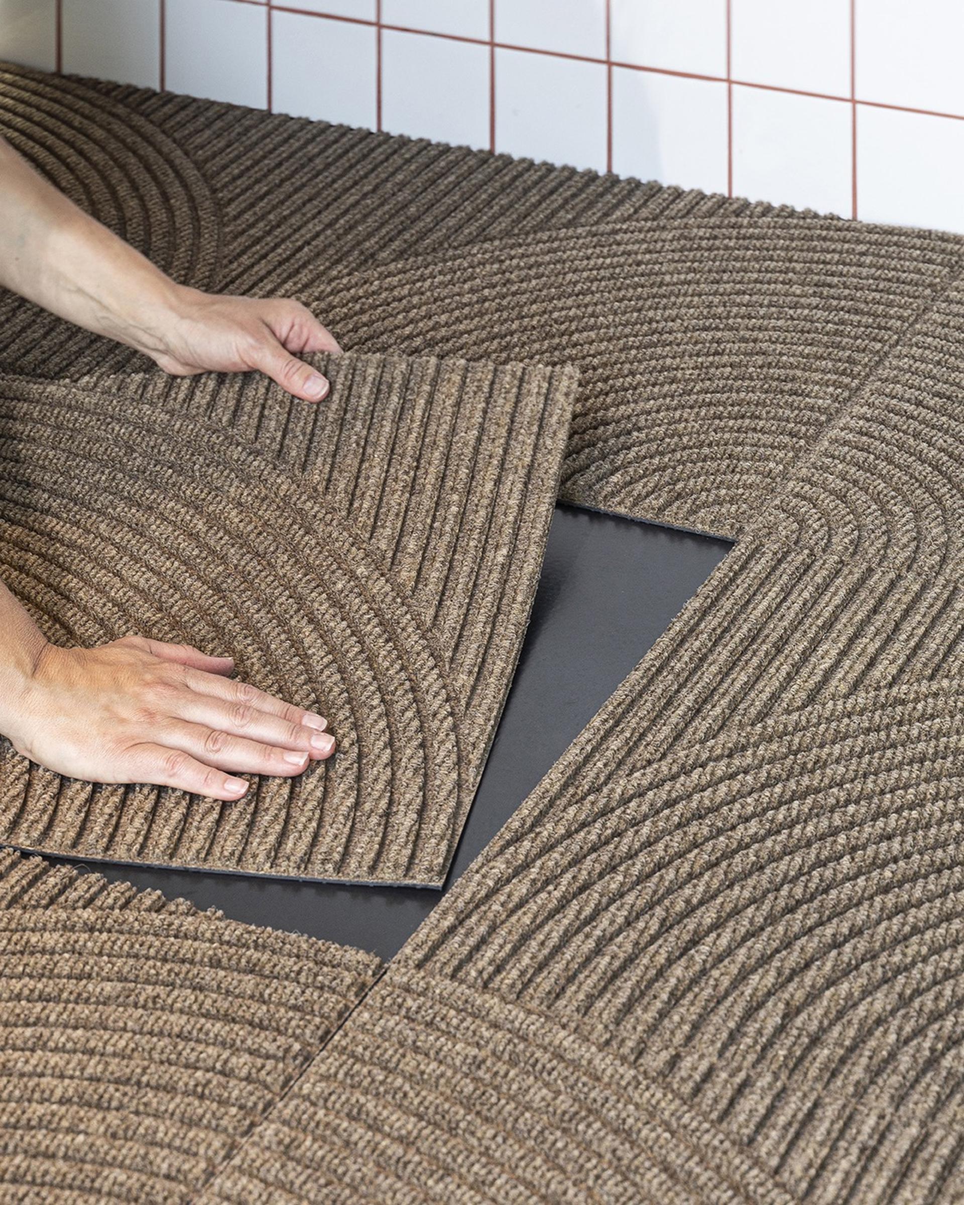 Hands placing interlocking brown Heymat mat tiles on a black surface with a red-tiled wall in the background.