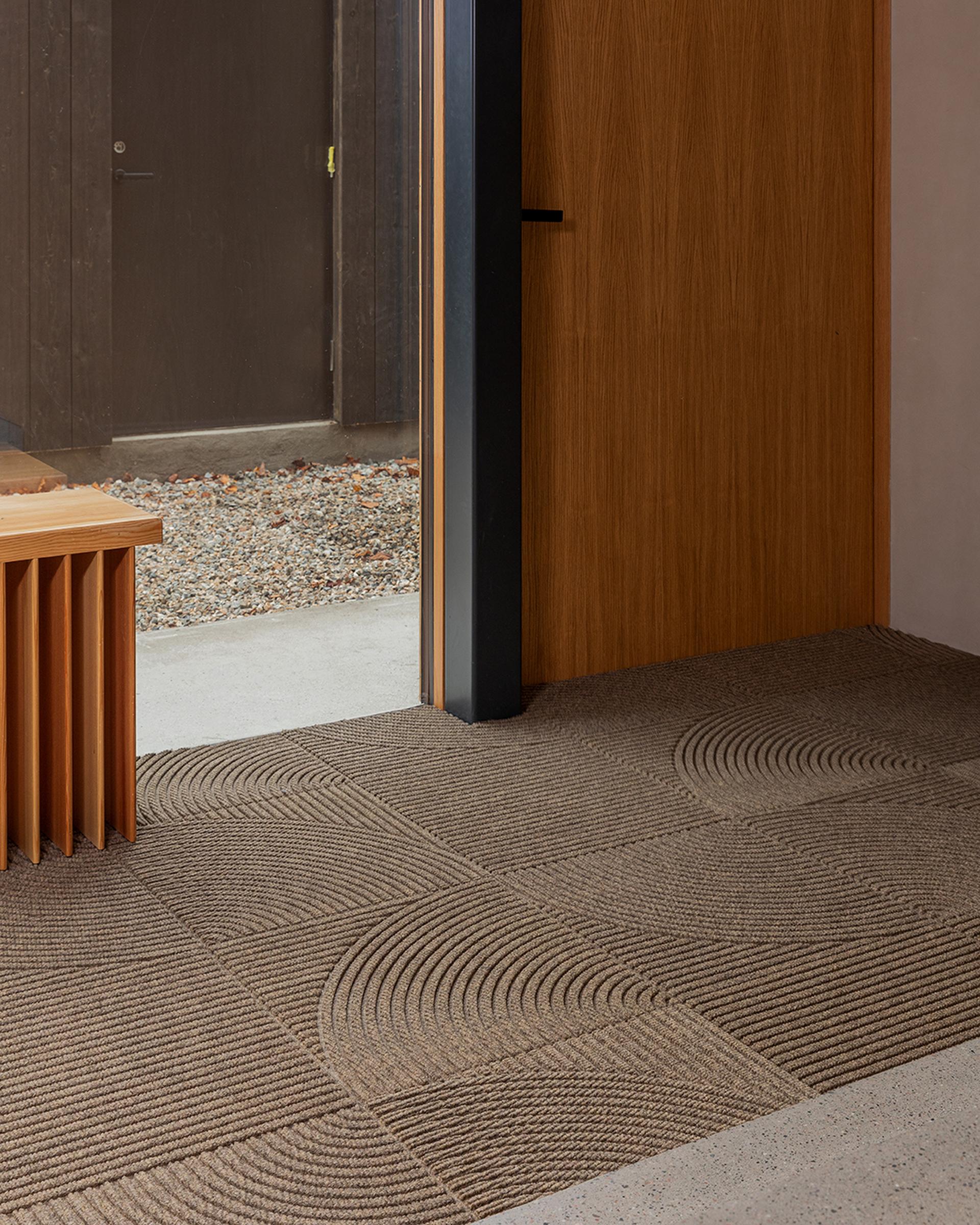 Modern interior with a wooden bench, brown textured carpet tiles by Heymat, and a partially open wooden door leading to a gravel path outside.