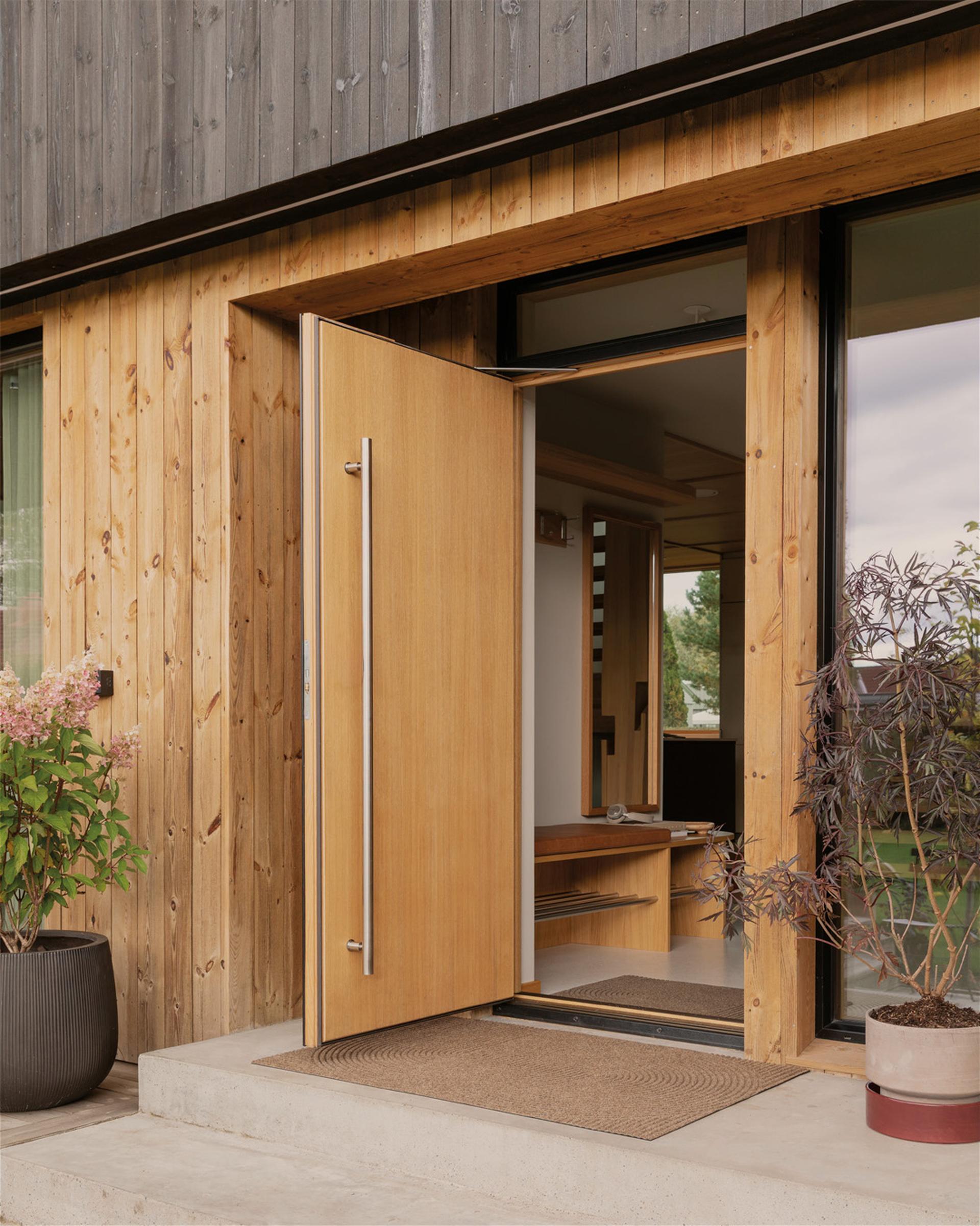 Wooden front door of a modern house, slightly ajar, with potted plants on either side and a doormat on a concrete step.