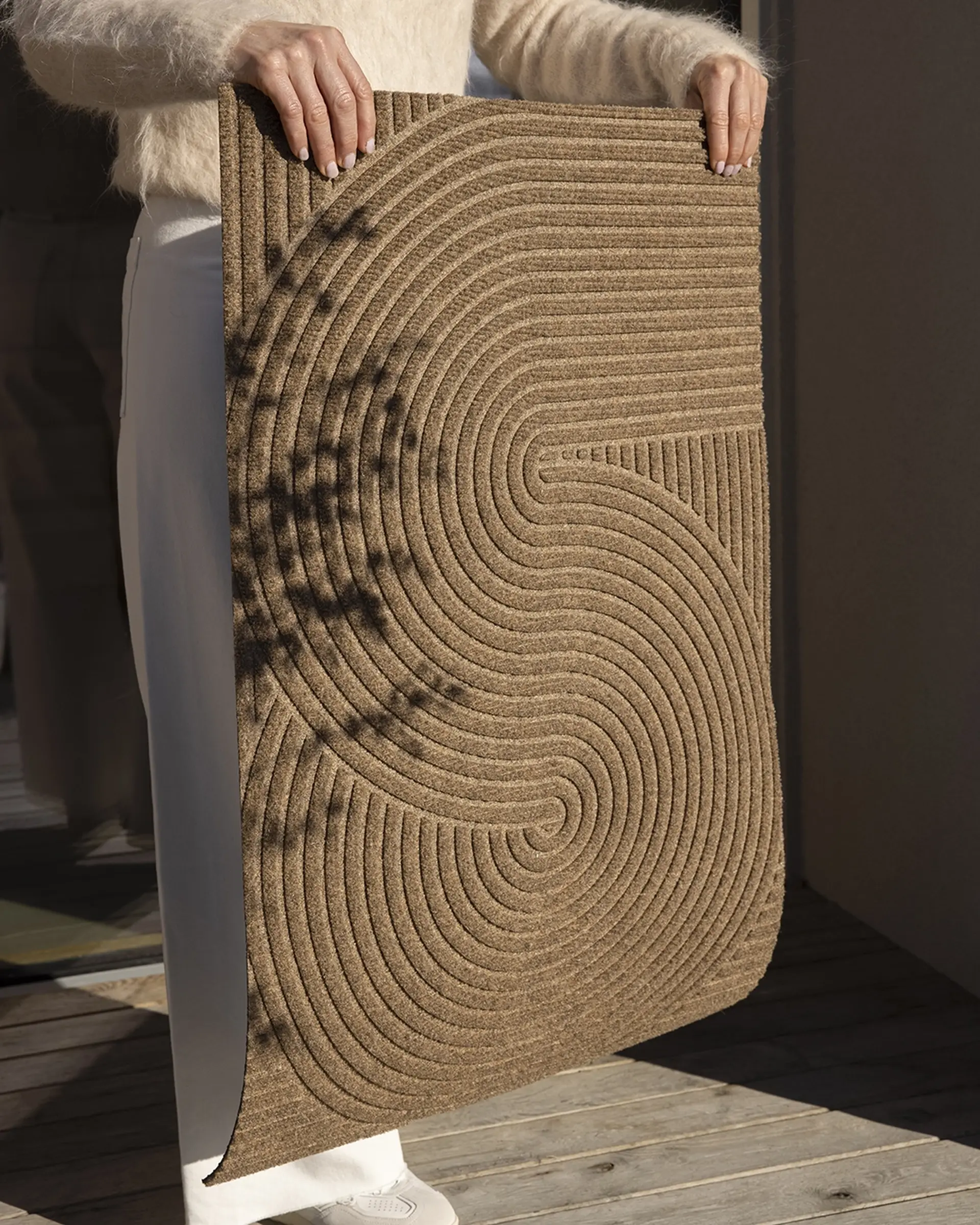 Person holding a textured brown doormat with intricate concentric swirl patterns, standing on a wooden deck.