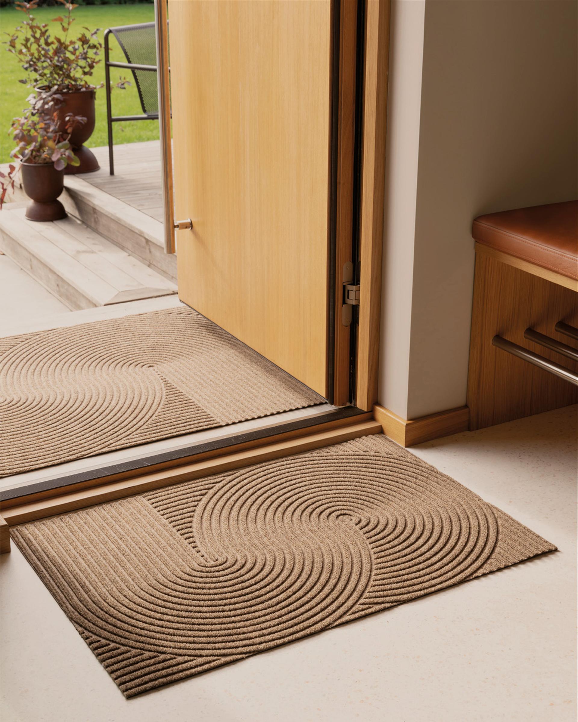 Entryway of a modern house, slightly ajar, with potted plants outside and two doormats on a concrete step.