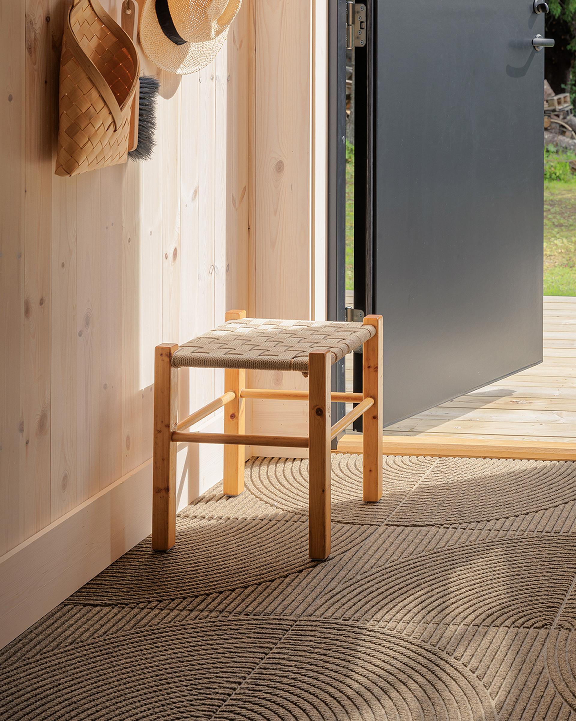 Sunlit wooden stool by an open door, casting shadows on textured Heymat Entry Mat Tiles. Woven basket and straw hat hang on a light wooden wall.