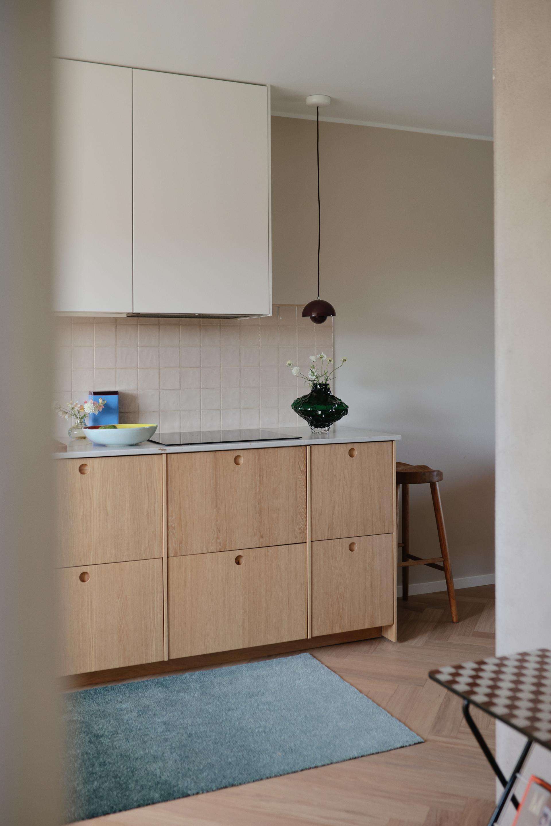 Modern kitchen with wooden cabinets, white countertops, green vase, blue bowl, and pendant light. Blue rug on wooden floor.