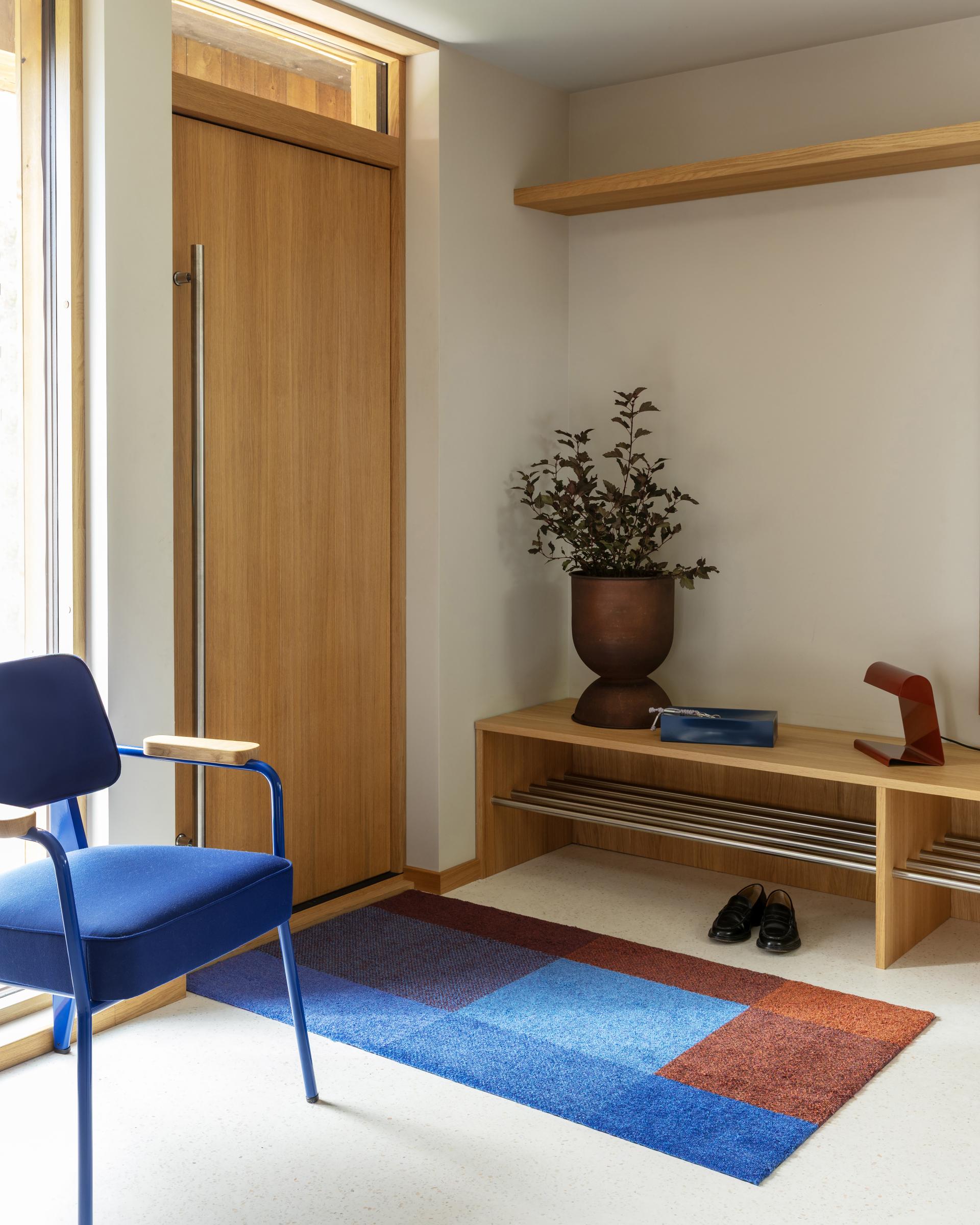 Modern entryway with wooden door, colorful geometric Mix Dusk doormat, blue chair, potted plant, and bench with shoes and decorative items.