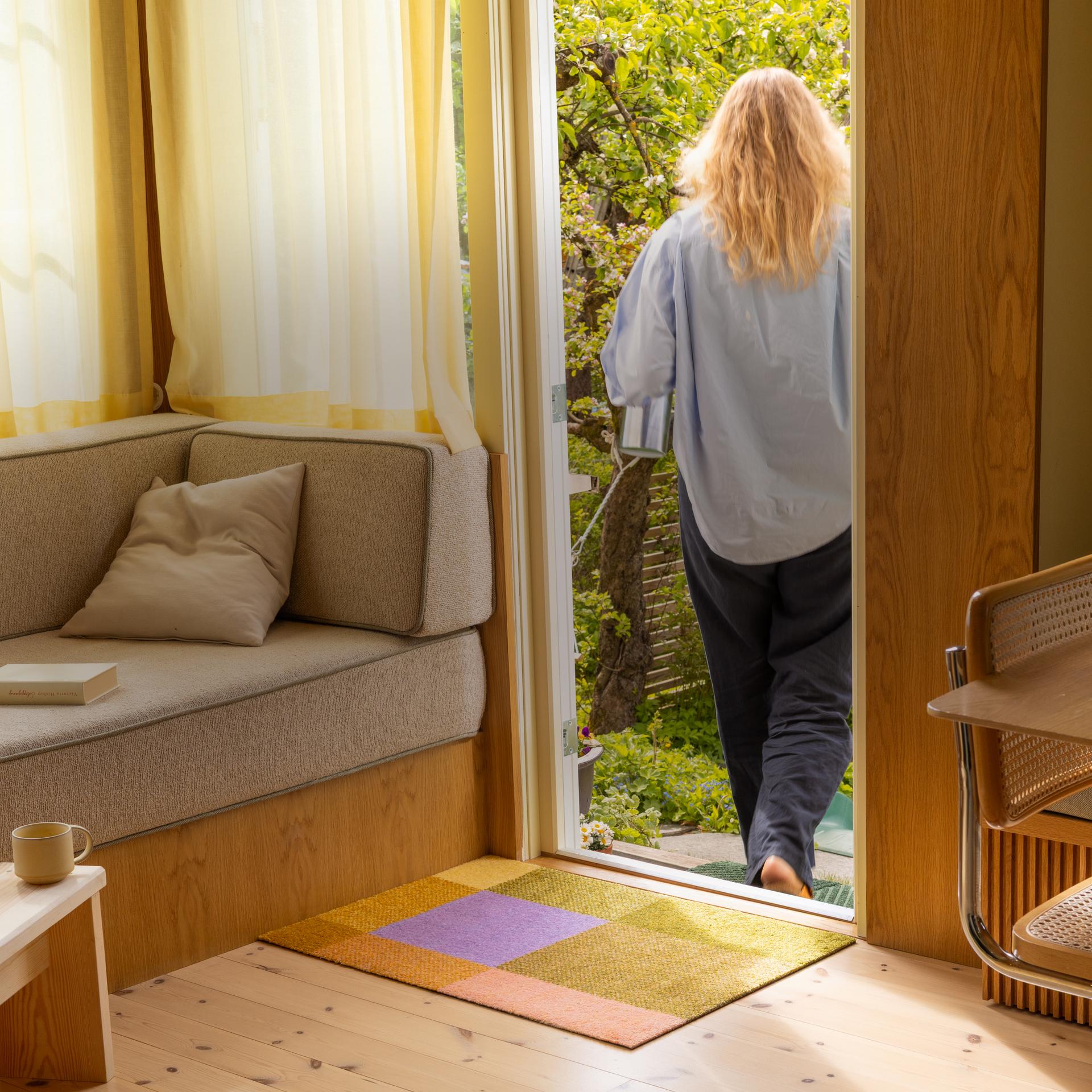 Person walking out of a cozy room with a Heymat Mix Teklan doormat onto a garden patio, with a sofa, table, and chairs inside. Sunlight filters through sheer curtains.