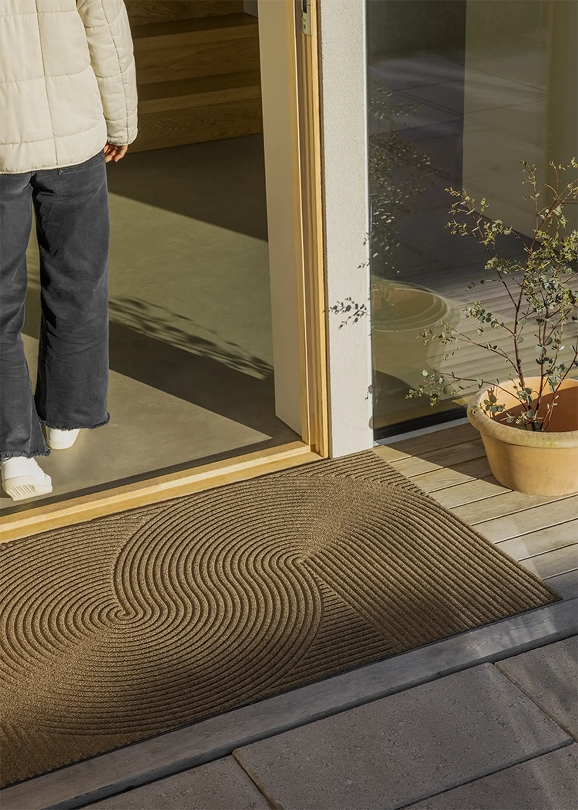 A person in a quilted jacket and black pants stands on a textured outdoor mat at an open doorway, with a potted plant nearby.