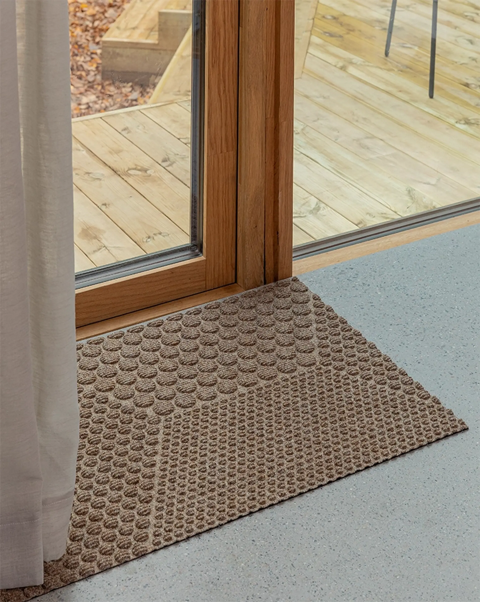 A brown textured doormat lies on a gray floor beside a glass door with a wooden frame, leading to an outdoor wooden deck.