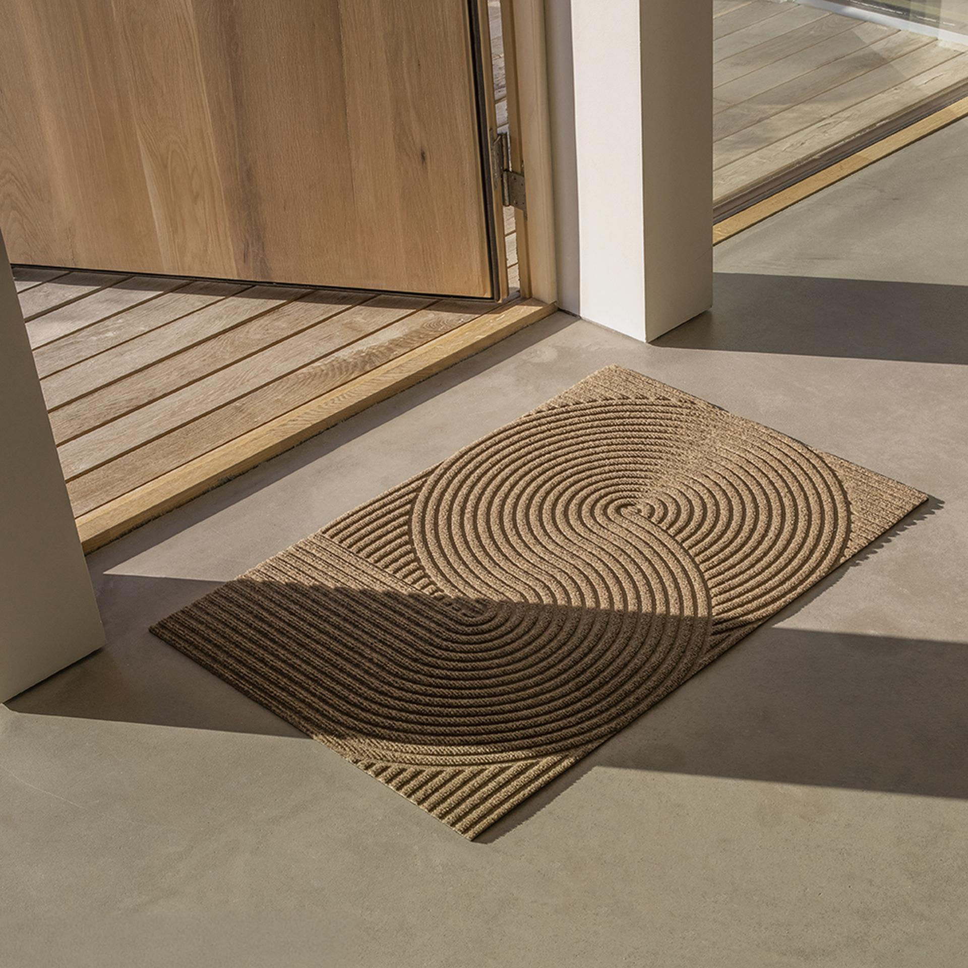 Heymat doormat with circular patterns on a concrete floor near an open wooden door, with sunlight casting shadows.
