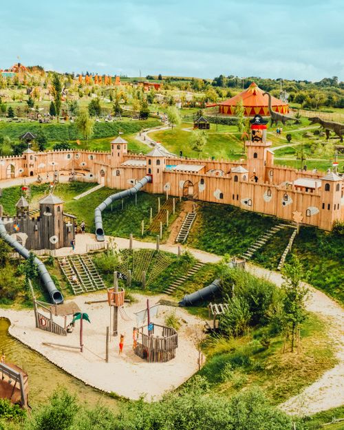 Spielplatz mit einer Festung im Piratenstil, Rutschen und Klettergerüsten, gelegen in einem grünen Park mit Dinosaurierfiguren und einem Zirkuszelt.