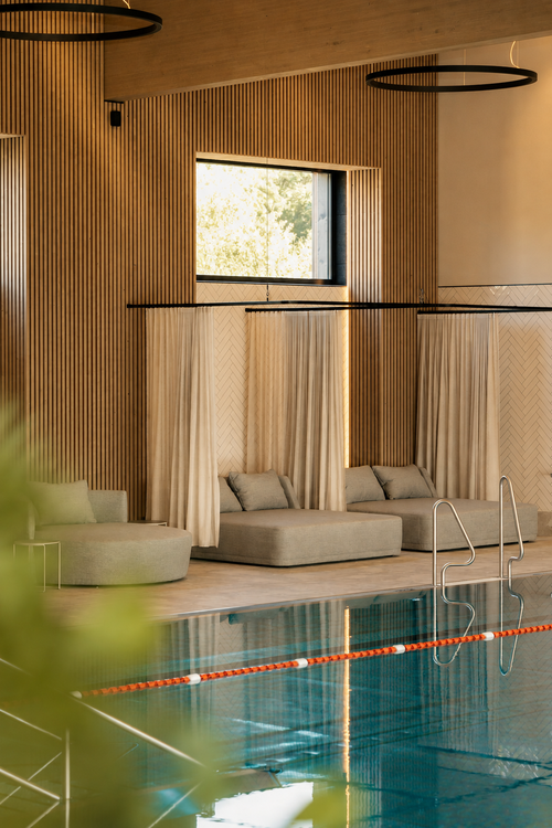 Indoorpool der IKUNA Spa- und Wellnesswelt mit gepolsterten Liegen, hellen Vorhängen und Holzlamellen an den Wänden. Große Fenster und warme Beleuchtung prägen den Ruhebereich am Beckenrand.