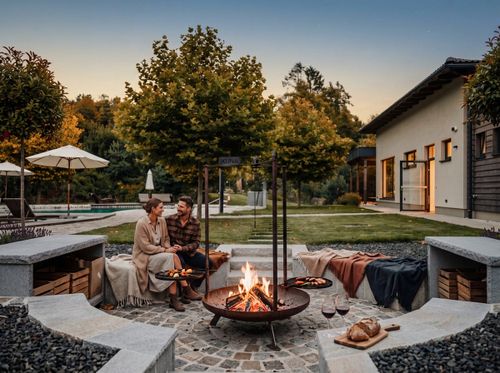 Ein Paar sitzt in der Abenddämmerung an einer Feuerstelle und grillt gemeinsam. Im Hintergrund befinden sich ein moderner Garten mit Bäumen und ein Wohnhaus.