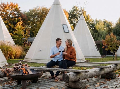 Ein Paar sitzt mit Weingläsern auf einer rustikalen Holzbank an einer Feuerstelle. Im Hintergrund stehen mehrere Tipis, umgeben von herbstlich gefärbten Bäumen.