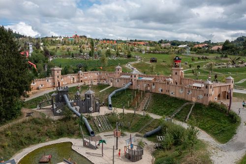 Piratenküste Spielplatz in Form einer Burg. Kinder rutschen und spielen. Die Umgebung ist grün mit Hügeln und Wegen im Hintergrund.