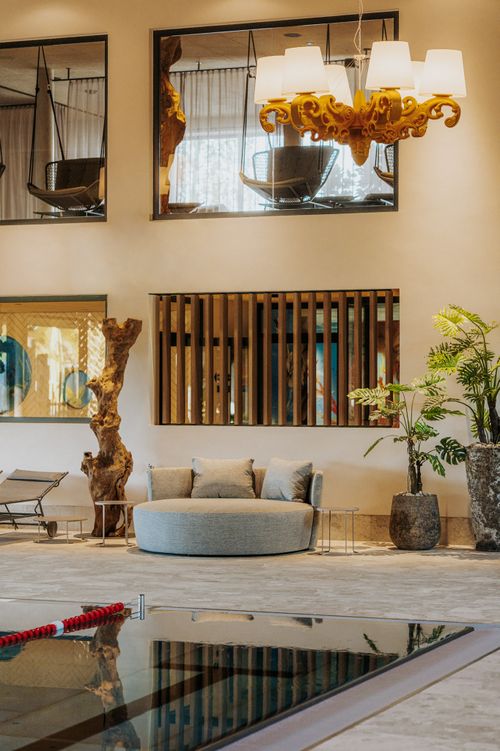 Rundes Daybed mit Kissen am IKUNA Indoorpool, umgeben von Pflanzen und Holzdekoration. Darüber hängt ein goldfarbener Kronleuchter; durch die Fenster sind Hängesessel im oberen Ruhebereich zu sehen.