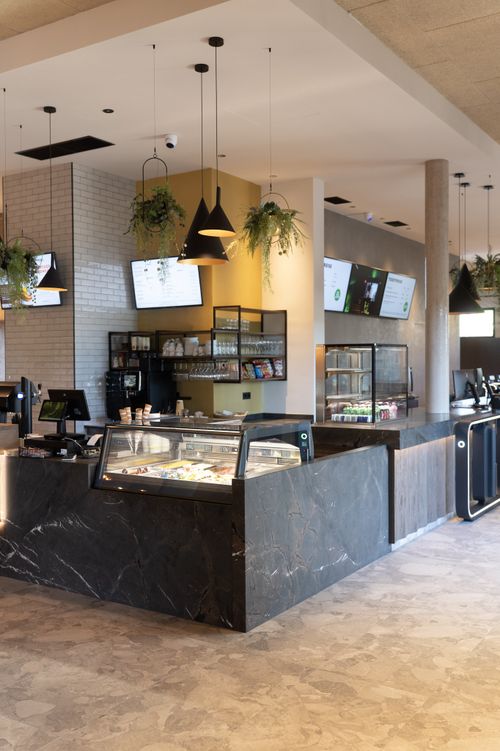 IKUNA Erlebnisbad Bistro Helles, modernes Café-Interieur mit Marmortheken, einer Konditorei-Vitrine, hängenden Pflanzen, Pendelleuchten und digitalen Menübildschirmen; warme, einladende Atmosphäre im IKUNA Erlebnisbad.
