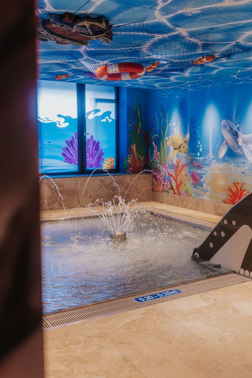 Flaches Kinderbecken mit Wasserfontäne und kleiner Rutsche in der IKUNA Spa- & Wellnesswelt. Bunte Unterwassermotive mit Schildkröte, Wal und Korallen schmücken die Wände.