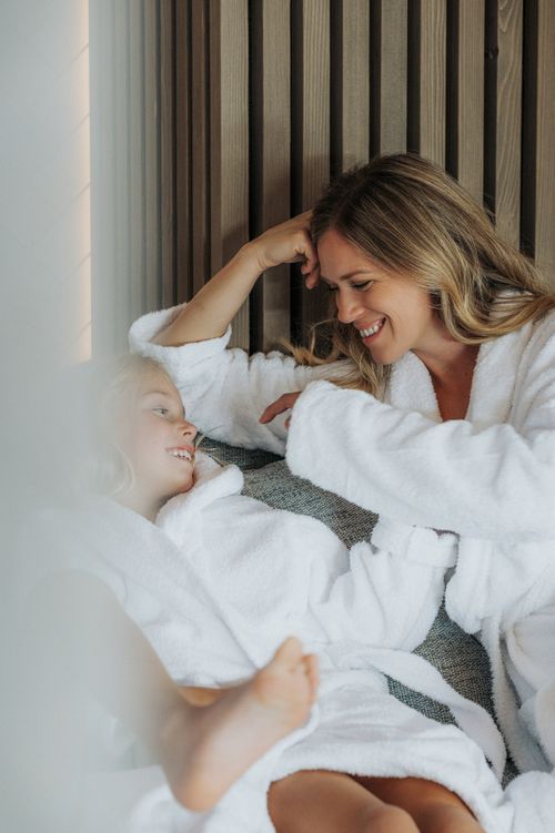 Eine Frau und ein Kind in weißen Bademänteln entspannen gemeinsam auf einer Liege in der IKUNA Spa- und Wellnesswelt und lächeln sich an. Im Hintergrund ist eine Wand aus Holzlamellen zu sehen.