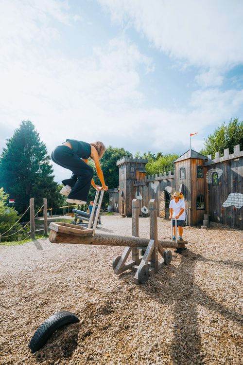 Ein Kind springt auf einer hölzernen Wippe auf einem Spielplatz im IKUNA Naturresort in Österreichs größen Piratenküste, im Hintergrund ist eine Spielanlage im Burgstil zu sehen. Die Szene strahlt Begeisterung und Verspieltheit aus.