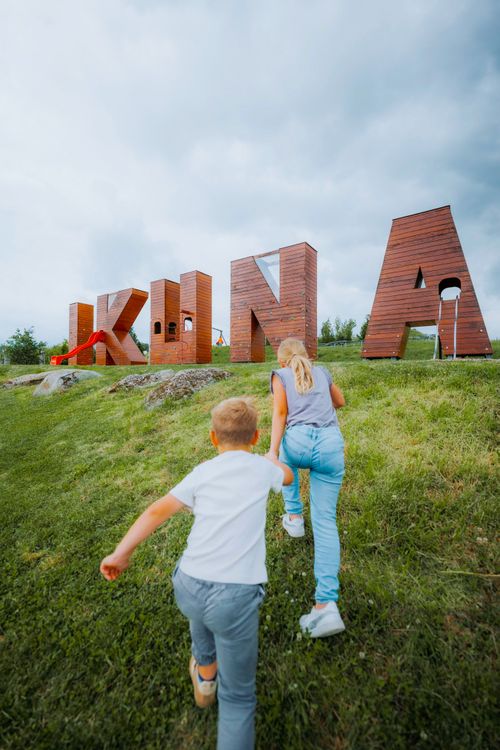 IKUNA Naturresort Zwei Kinder laufen einen grasbewachsenen Hügel hinauf in Richtung großer Holzbuchstaben, die das Wort „IKUNA“ bilden. In einen der Buchstaben ist eine rote Rutsche eingebaut, über ihnen erstreckt sich ein bewölkter Himmel. Es herrscht eine ausgelassene Atmosphäre im IKUNA Naturresort.