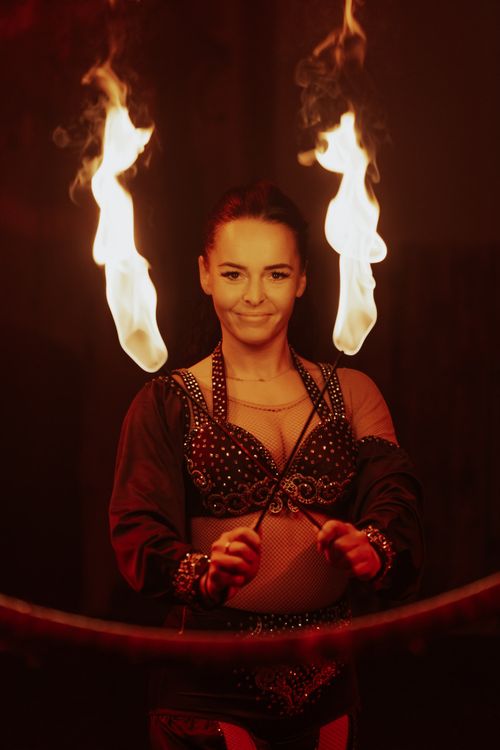 Eine Person jongliert zwei Feuerfackeln in bunter Kleidung, umgeben von dunkler, stimmungsvoller Atmosphäre.