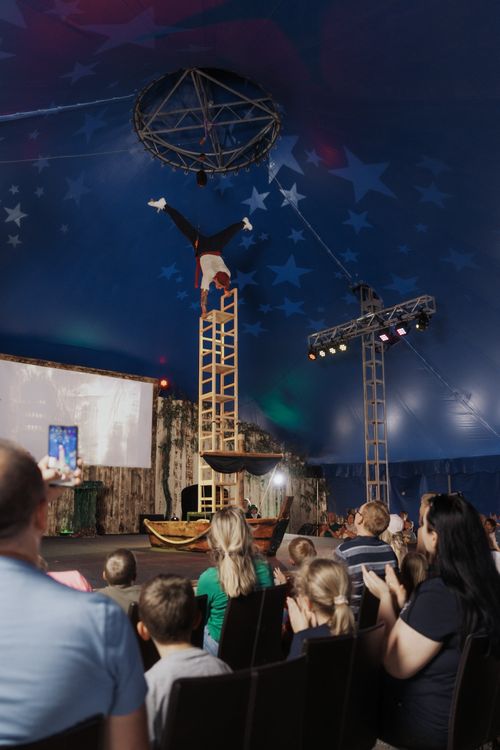 IKUNA Zirkusshow Akrobat balanciert kopfüber auf mehreren Stühlen, in einem Zirkuszelt. Zuschauer applaudieren, während ein Bildschirm im Hintergrund sichtbar ist.