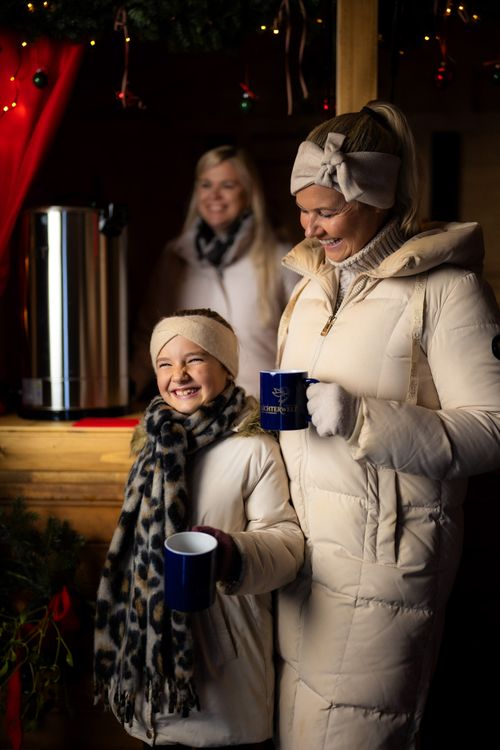 Eine Mutter und ihre lächelnde Tochter in Wintermänteln halten an einem festlich geschmückten Weihnachtsstand in der IKUNA Lichterwelt, umgeben von funkelnden Lichtern und einer Frau hinter ihnen, Tassen mit Kakao in den Händen.
