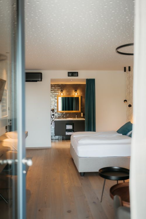 Zimmer enthält ein Bett und Sofa, mit einem Badezimmer im Hintergrund. Moderne Einrichtung, Holzfußboden, weiche Beleuchtung und dezentes Design.