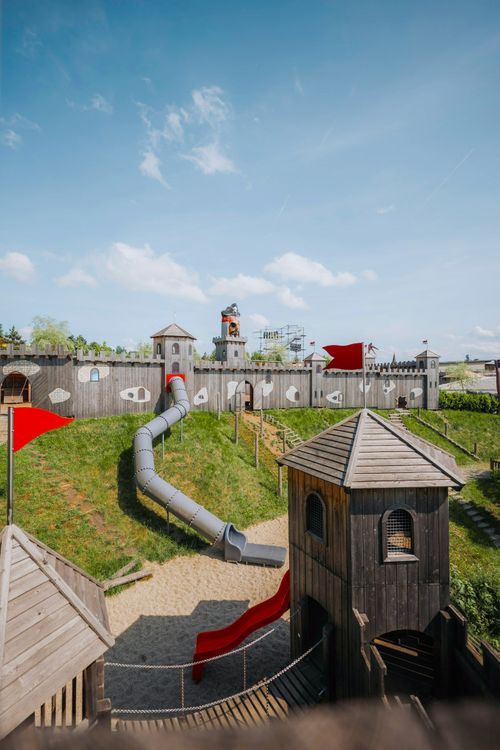 IKUNA Naturresort Dřevěné hřiště ve tvaru hradu s věžemi a skluzavkami. Zářivě červené vlajky a jasně modrá obloha navozují v IKUNA Naturresortu hravou a slunnou atmosféru.