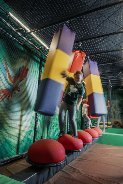 Kind balanciert auf roten Kugeln, umgeben von großen, bunten Hindernissen in einer Indoor-Spielanlage mit Dschungelmotiven an den Wänden.