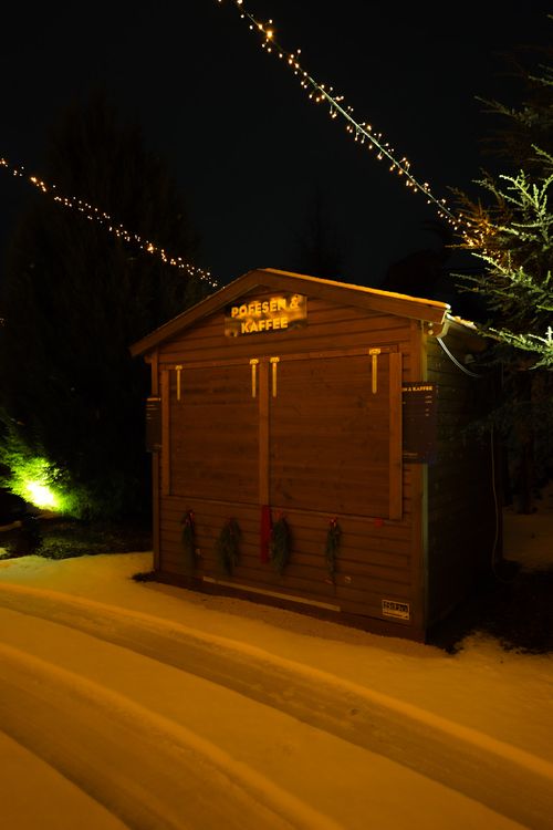 Ein schneebedeckter hölzerner Punschhütte bei Nacht, warm beleuchtet und mit Lichterketten geschmückt, der vor dem Hintergrund dunkler immergrüner Bäume leuchtet; gemütliche, festliche Stimmung in der IKUNA Lichterwelt.