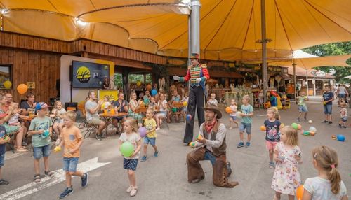 Eine lebhafte Festivalstimmung in der IKUNA City, mit Kindern, die bunte Luftballons in den Händen halten. Ein Stelzenläufer und ein Cowboy-Jongleur sorgen unter großen gelben Markisen für Unterhaltung.