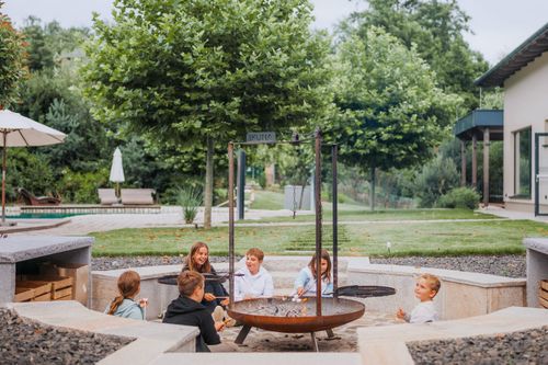 IKUNA Naturresort Děti sedící kolem ohniště a opékající marshmallows na zahradě obklopené zelenými stromy a s bazénem v blízkosti vytvářejí v IKUNA Naturresort radostnou atmosféru.