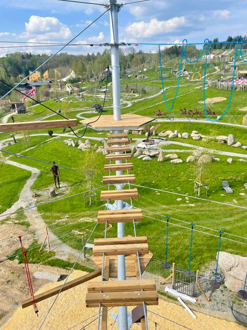 IKUNA Naturresort Vysoká lanová dráha v zábavním parku s dřevěnými plošinami a lany, zasazená do svěží zelené krajiny pod částečně zataženou oblohou v IKUNA Naturresort