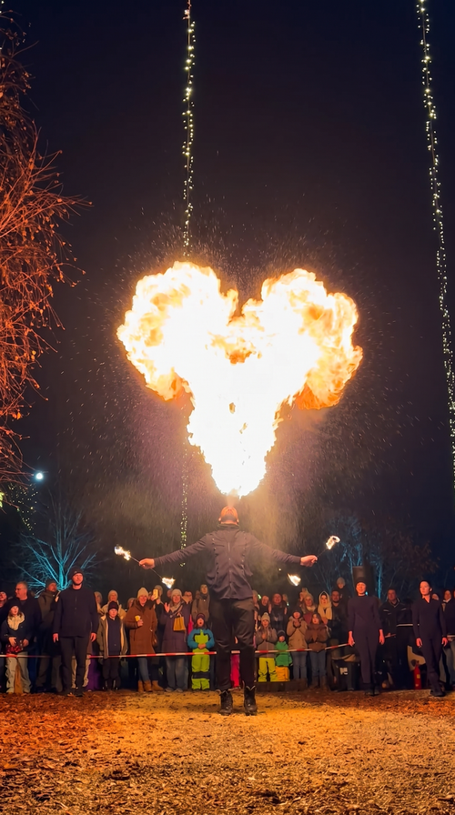 Bei der IKUNA Lichterwelt breitet ein Artist unter einer riesigen, herzförmigen Flamme, die aus seinem Mund kommt, die Arme aus, während eine dicht gedrängte Menschenmenge voller Ehrfurcht zusieht.
