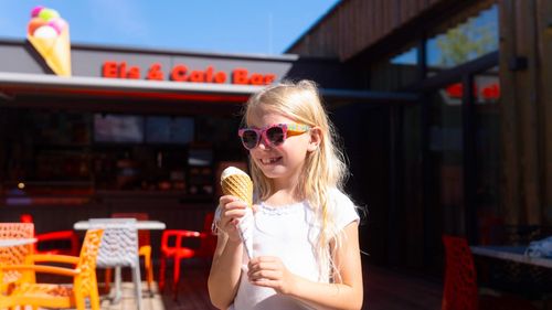 IKUNA_Zeugnis-Eisaktion-Keyvisual Blondes Mädchen steht mit Eis vor der IKUNA Eisbar im abenteuerlichen Naturerlebnispark.
